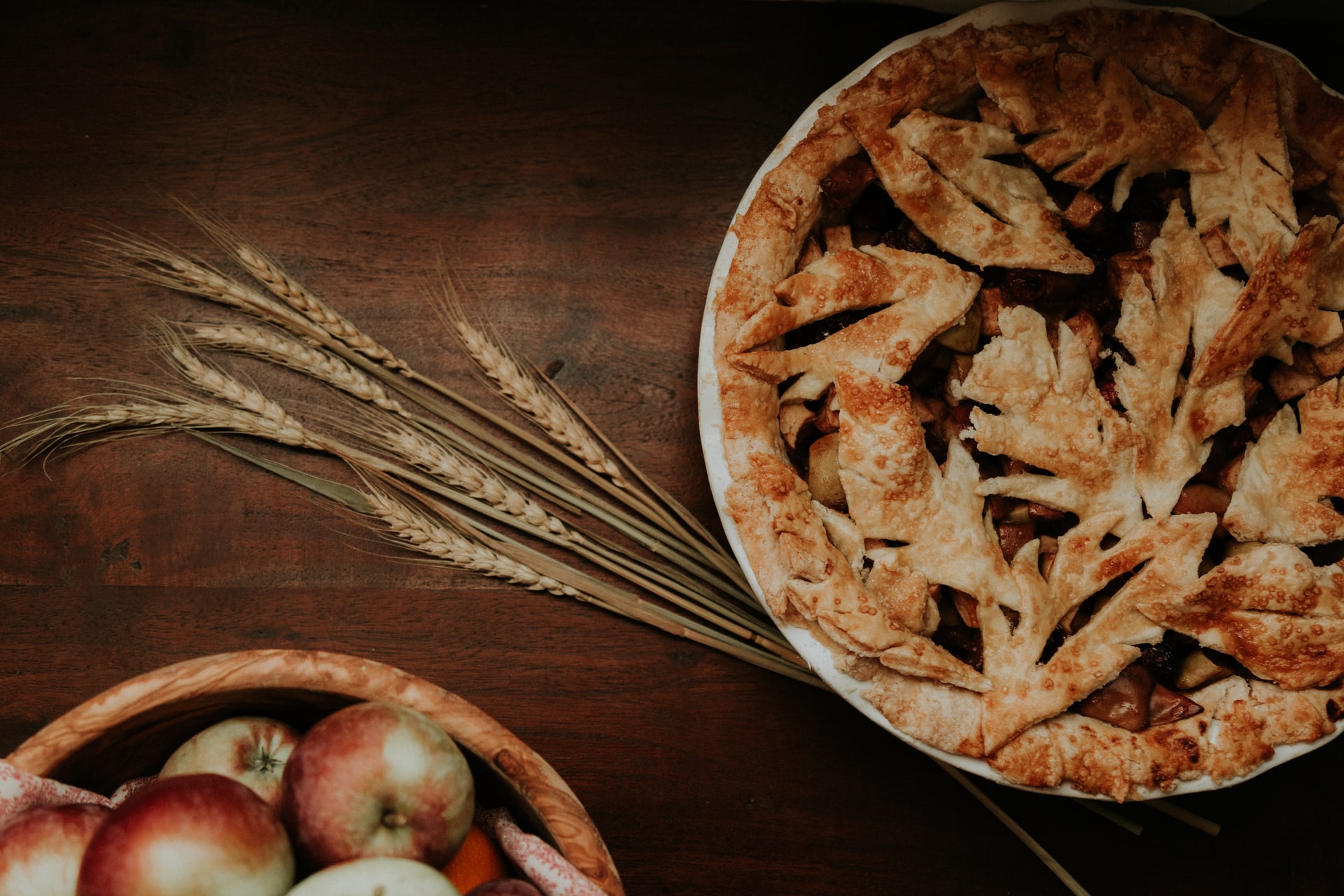 baked apple pie next to strands of wheat and a bowl of apples