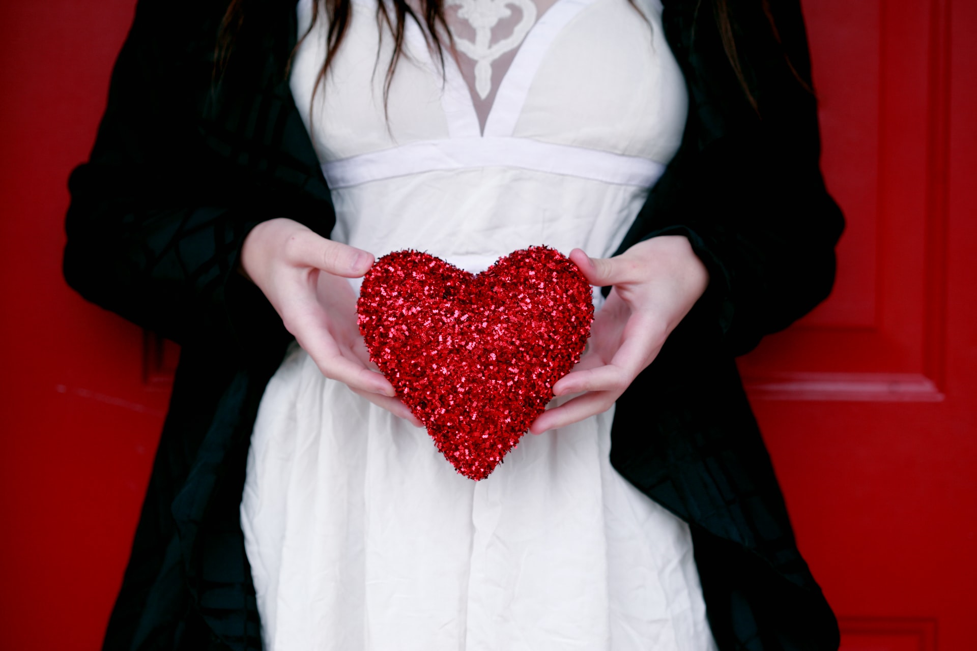 woman in a white dress and black coat holding a red glitter heart against a red background