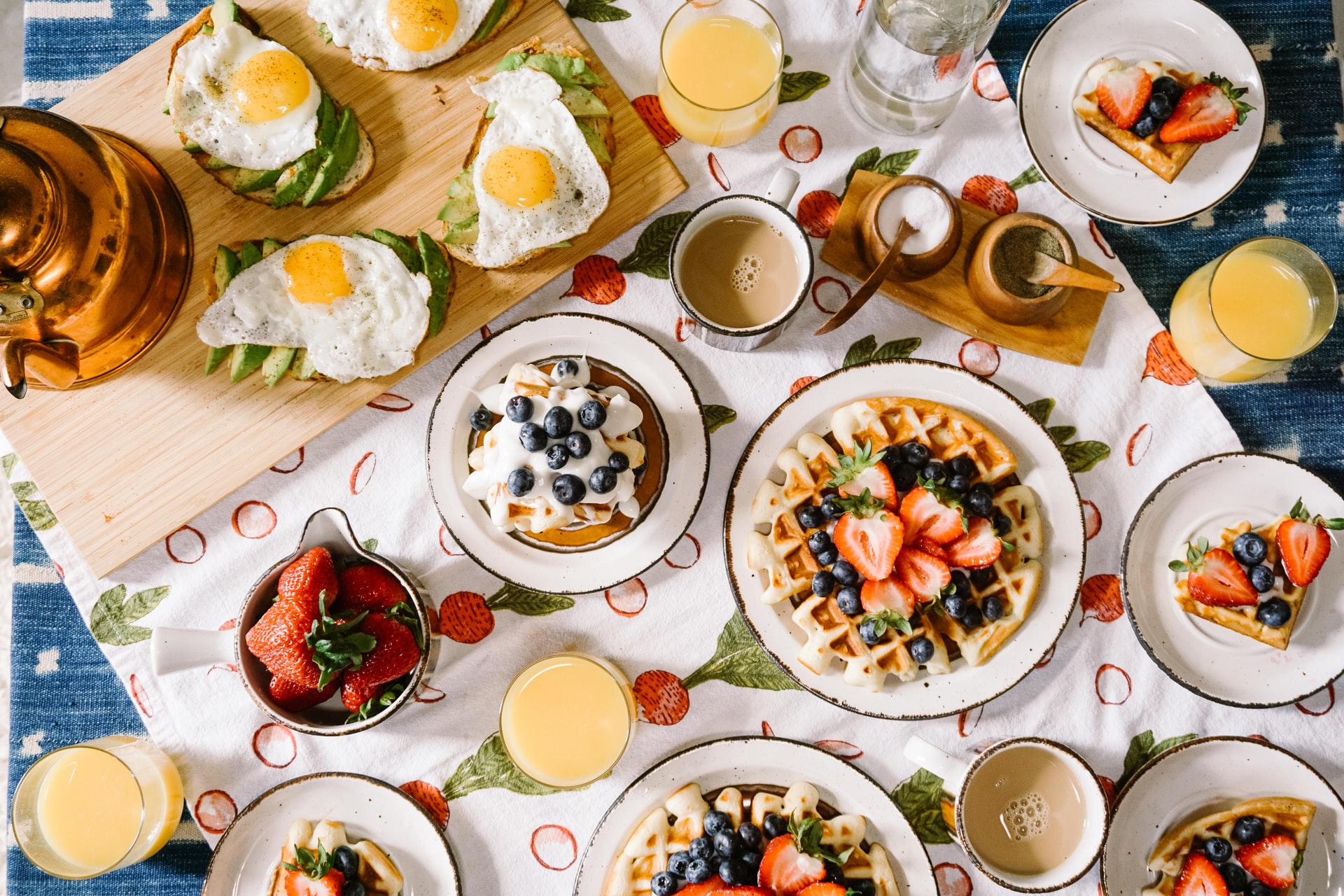 table filled with fruit waffles and pancakes and a platter of avocado toast topped with fried eggs