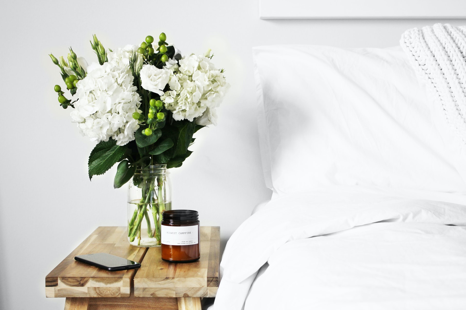 white bed with a wooden nightstand next to it containing a bouquet of white flowers, a candle and a cellphone