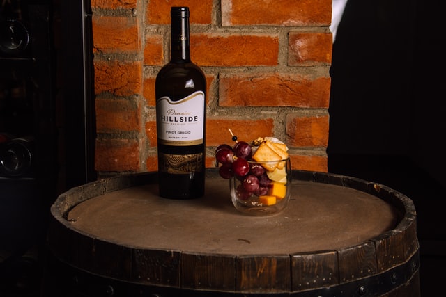 bottle of wine next to a cup containing fruit, cheese, and crackers on a barrel