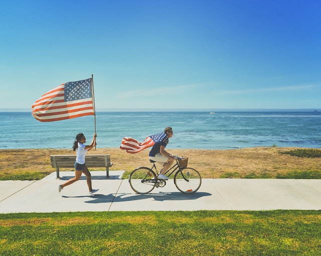 person on bicycle wearing USA flag as a cape, person following on foot waving USA flag on pole